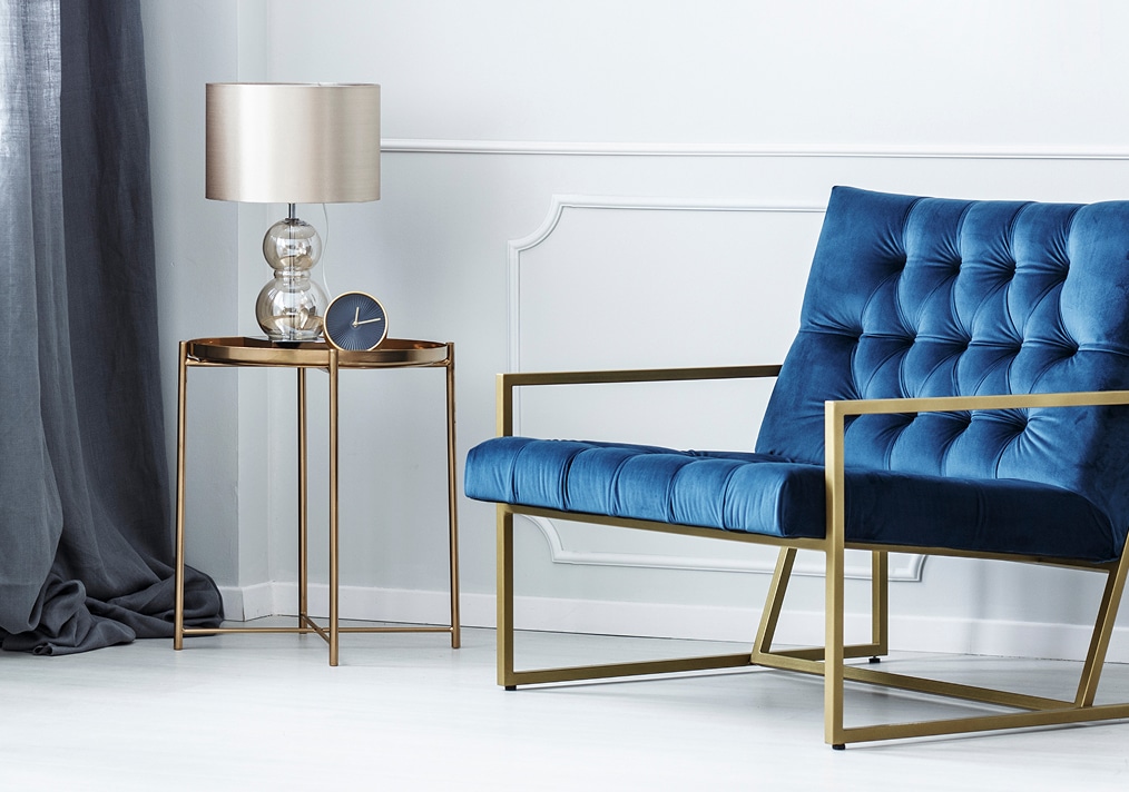 Fauteuil velours bleu & structure dorée | Déco moderne Fauteuil bas en velours bleu capitonné sur structure dorée, à côté d'une table d'appoint en métal contenant une lampe et une horloge design.