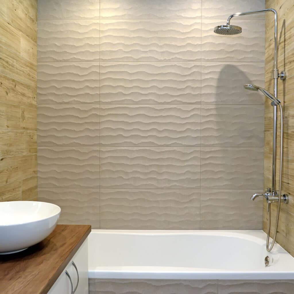 Carrelage vague et bois pour salle de bain design avec baignoire. Baignoire encastrée sous un mur de carrelage beige texturé effet vague. Côtés en carrelage imitation bois et vasque sur meuble.