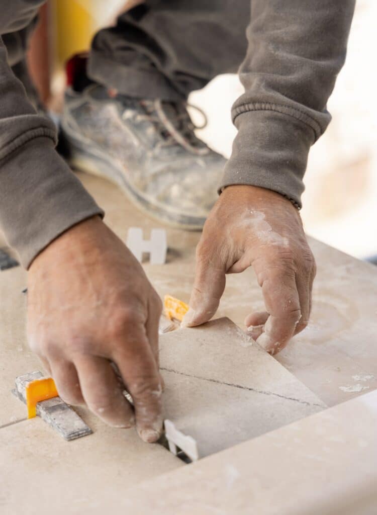 Pose de carrelage professionnelle : Nivellement parfait des carreaux. Gros plan des mains d'un carreleur ajustant un carreau avec des clips de nivellement. Poussière de ciment sur les mains et les bottes.
