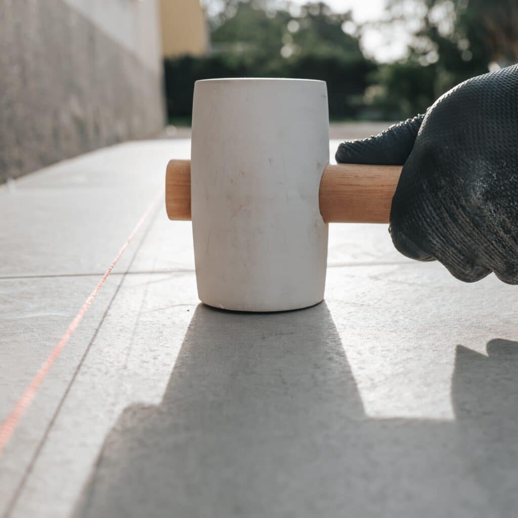 Pose de carrelage extérieur : ajuster les dalles au maillet Main gantée utilisant un maillet en caoutchouc blanc pour fixer une dalle de carrelage extérieur, guidée par un cदेrdeau rouge.
